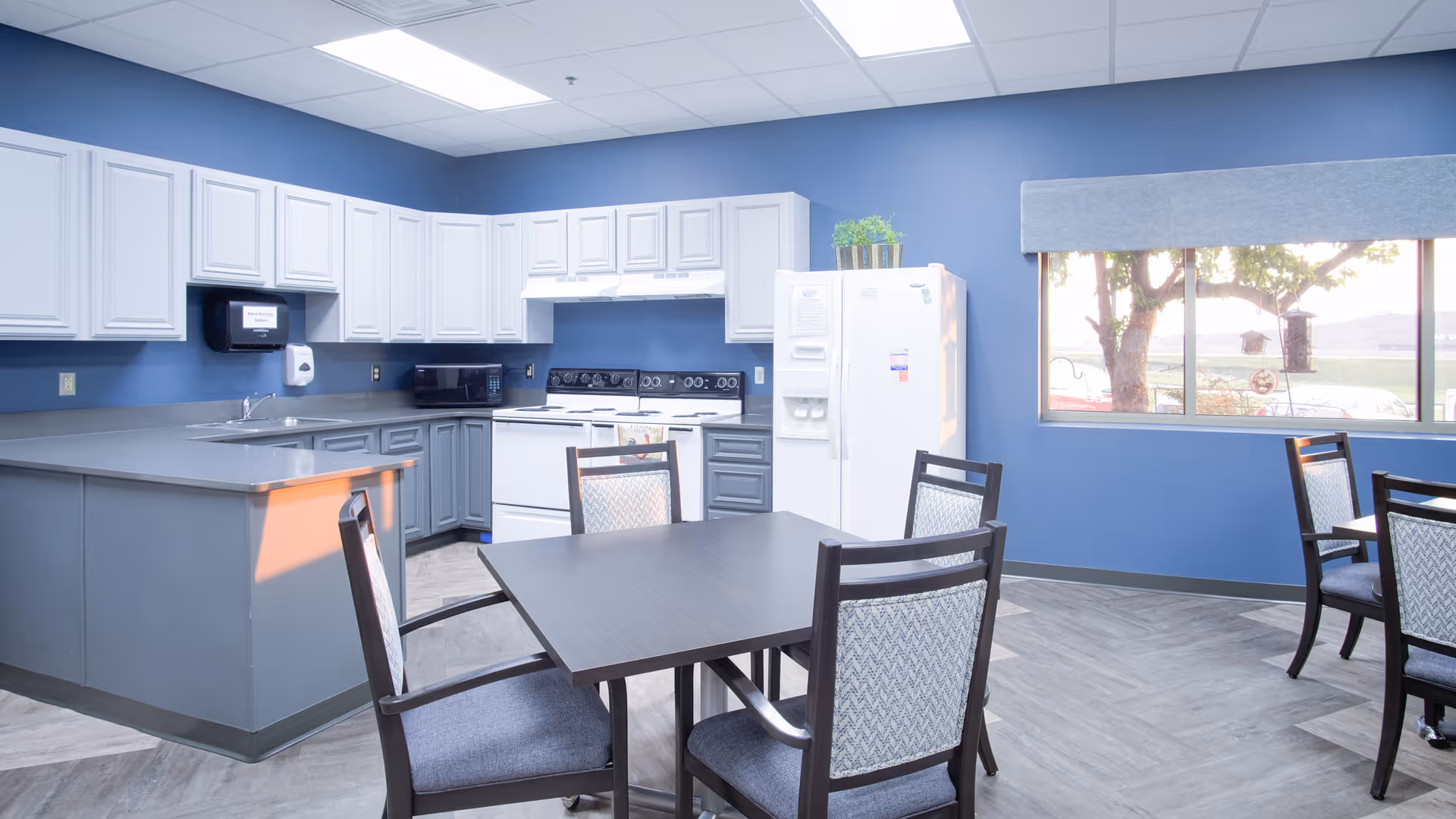A bright kitchen and dining area with blue walls, white upper cabinets, gray lower cabinets, a white stove, microwave, and refrigerator. There are two dark wood tables with four chairs each, and a large window with a gray valance letting in natural light.