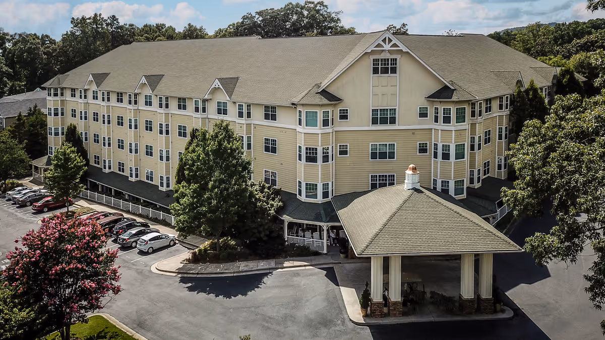 A large, multi-story senior living facility building with beige siding and numerous windows, surrounded by trees and a parking lot with cars. The entrance features a covered drop-off area with a peaked roof. The sky is partly cloudy.