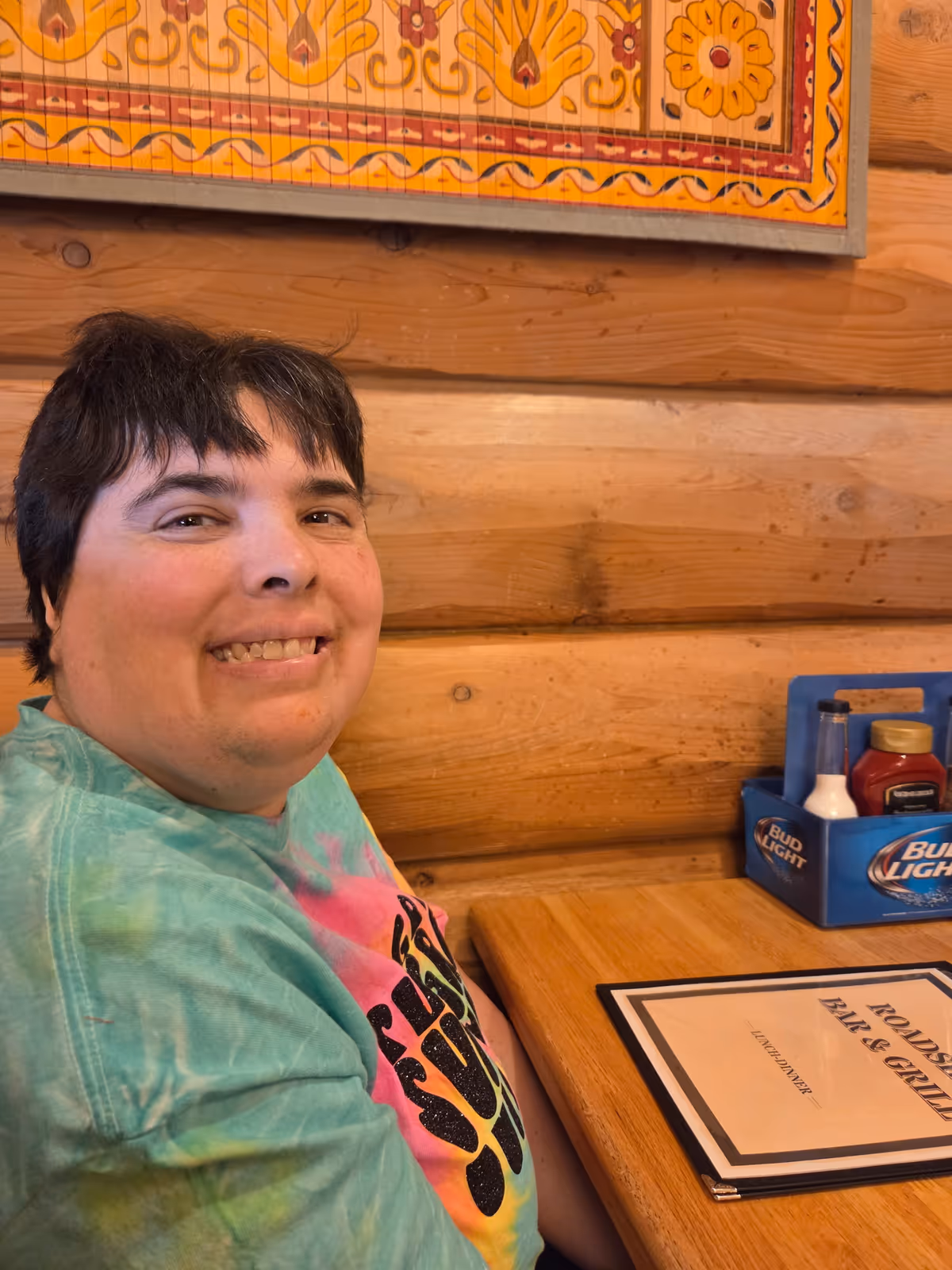 A person with short dark hair wearing a colorful tie-dye shirt is sitting at a wooden table in a rustic restaurant with log cabin style walls. On the table is a menu labeled 'Roadside Bar & Grill' and a blue container holding condiments including ketchup, mustard, and a bottle of white sauce. A decorative framed artwork with yellow and red patterns hangs on the wall behind.