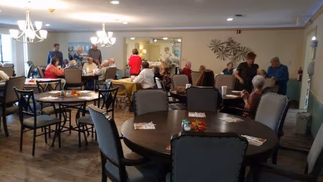 A dining room in an assisted living community with several elderly residents seated at round tables, engaging in conversation and activities. The room is well-lit with chandeliers and has a warm, inviting atmosphere with neutral-colored walls and wooden flooring.