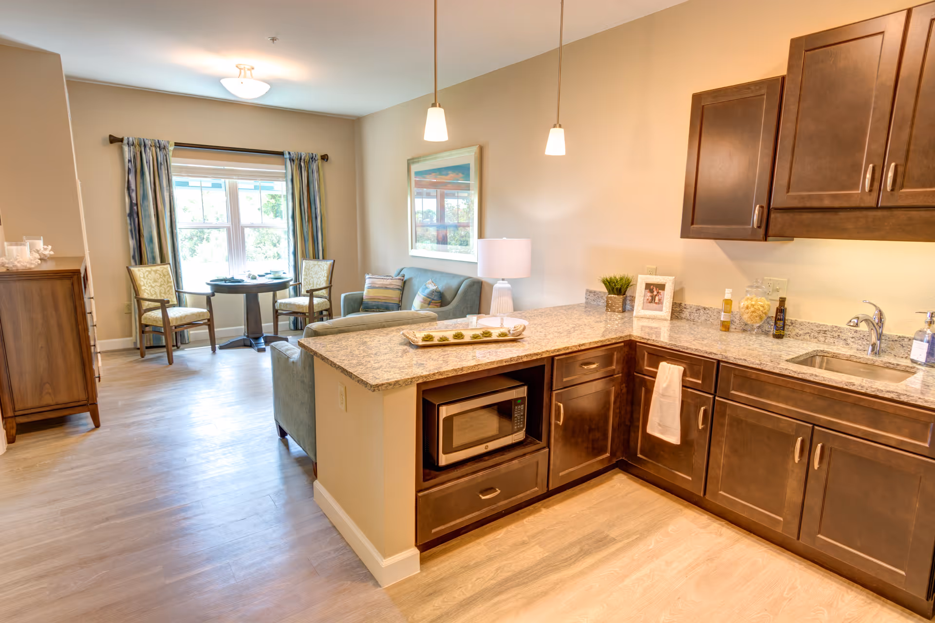 A bright and cozy living space featuring a kitchen area with dark wood cabinets, granite countertops, a microwave, and a sink. Adjacent to the kitchen is a small seating area with a light blue sofa, a table lamp, and a framed picture on the wall. In the background, there is a dining area with a round table and two chairs near a window with patterned curtains, allowing natural light to fill the room.
