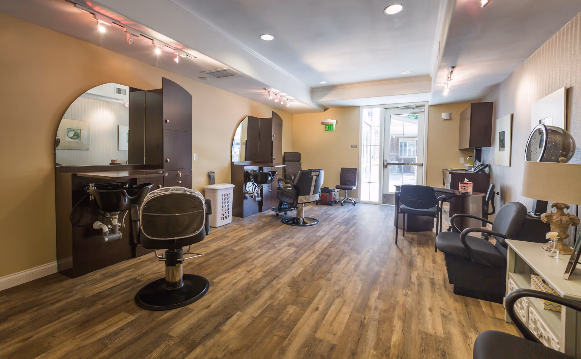 Interior view of a salon area in an assisted living facility featuring two styling stations with mirrors and chairs, a wooden floor, a seating area with chairs and a table, and a door leading outside.