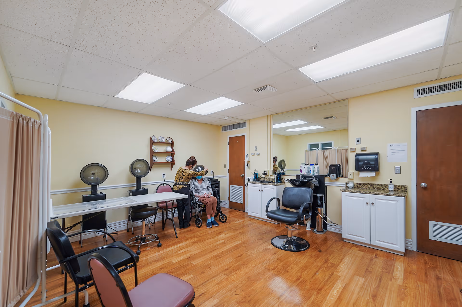 A hair salon room in a senior living facility with wooden floors, yellow walls, and fluorescent ceiling lights. There are salon chairs, hair dryers, a large mirror, and a counter with hair care products. A woman is styling the hair of an elderly person seated in a wheelchair.