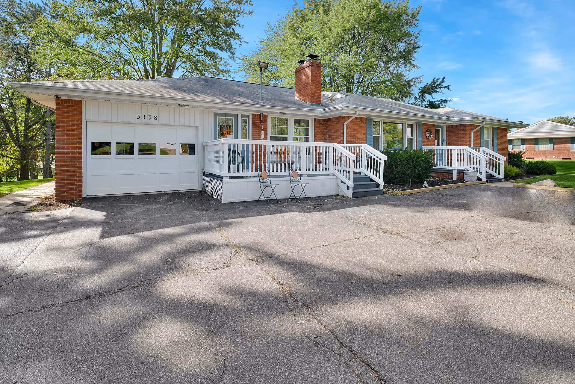 Exterior view of a single-story brick building with a white garage door and two white-railed porches. There are two chairs on the porch in front of the garage. The building is surrounded by trees and greenery under a clear blue sky.