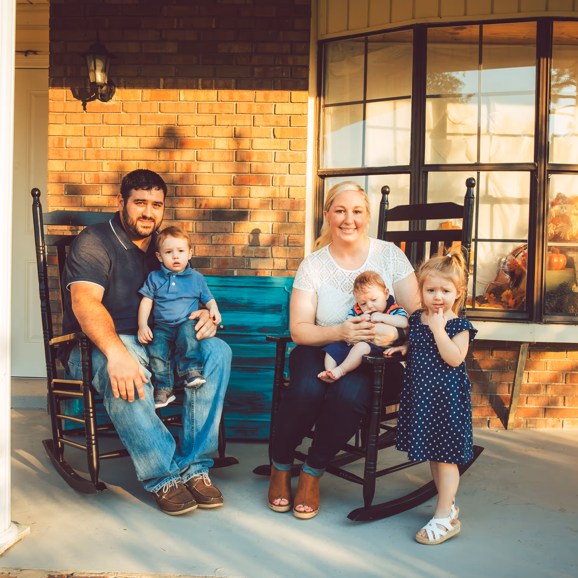 A family of five sitting and standing on a porch in front of a brick wall and window. A man is sitting on a rocking chair holding a young boy, a woman is sitting on another rocking chair holding a baby, and a young girl in a polka dot dress is standing nearby.