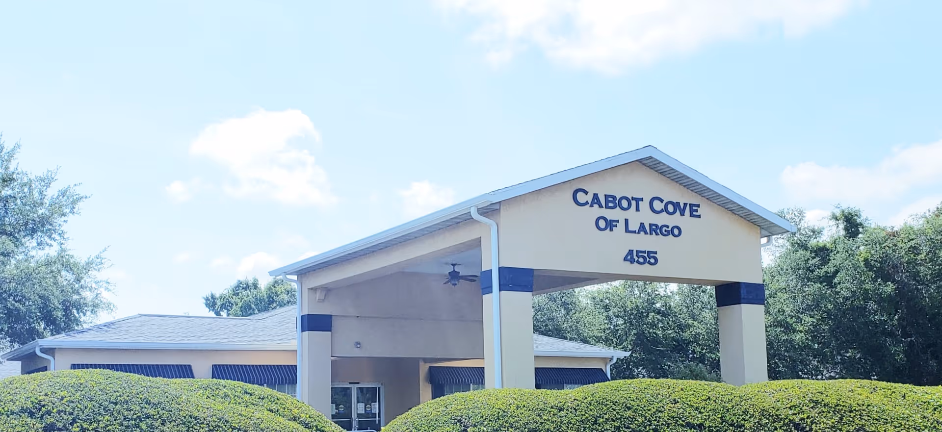 Exterior front view of Cabot Cove of Largo building entrance with a covered driveway, surrounded by green bushes and trees under a partly cloudy sky.