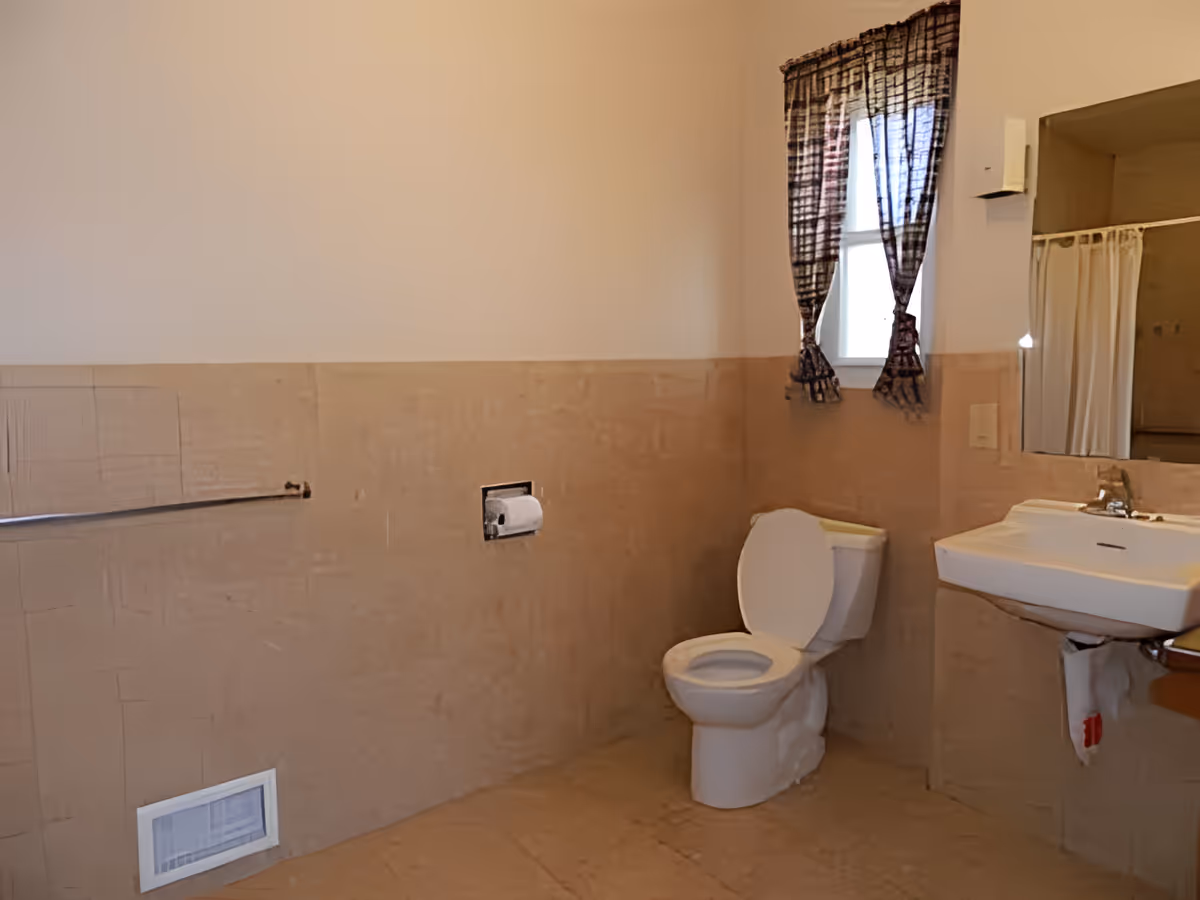 A bathroom with a toilet, a wall-mounted sink, a mirror, a window with dark patterned curtains, a towel bar, and a toilet paper holder. The walls are tiled halfway up and painted beige above the tiles.