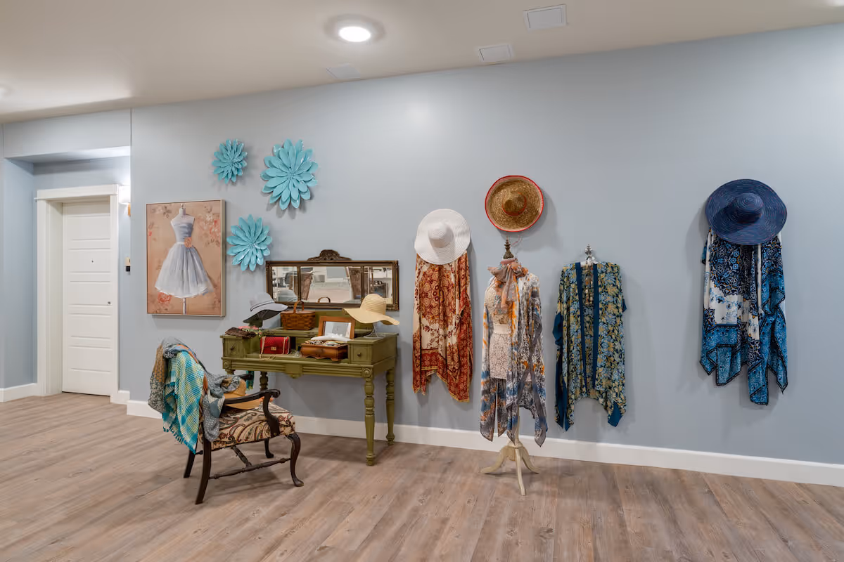 A stylish interior corner of a senior living facility with a light blue wall decorated with large blue flower wall hangings, a painting of a dress on a mannequin, and several hats and colorful scarves displayed on hooks and a mannequin. There is a vintage green desk with a mirror and various items on top, and an ornate wooden chair with a patterned cushion and a blue shawl draped over it. The floor is wood with a light finish.