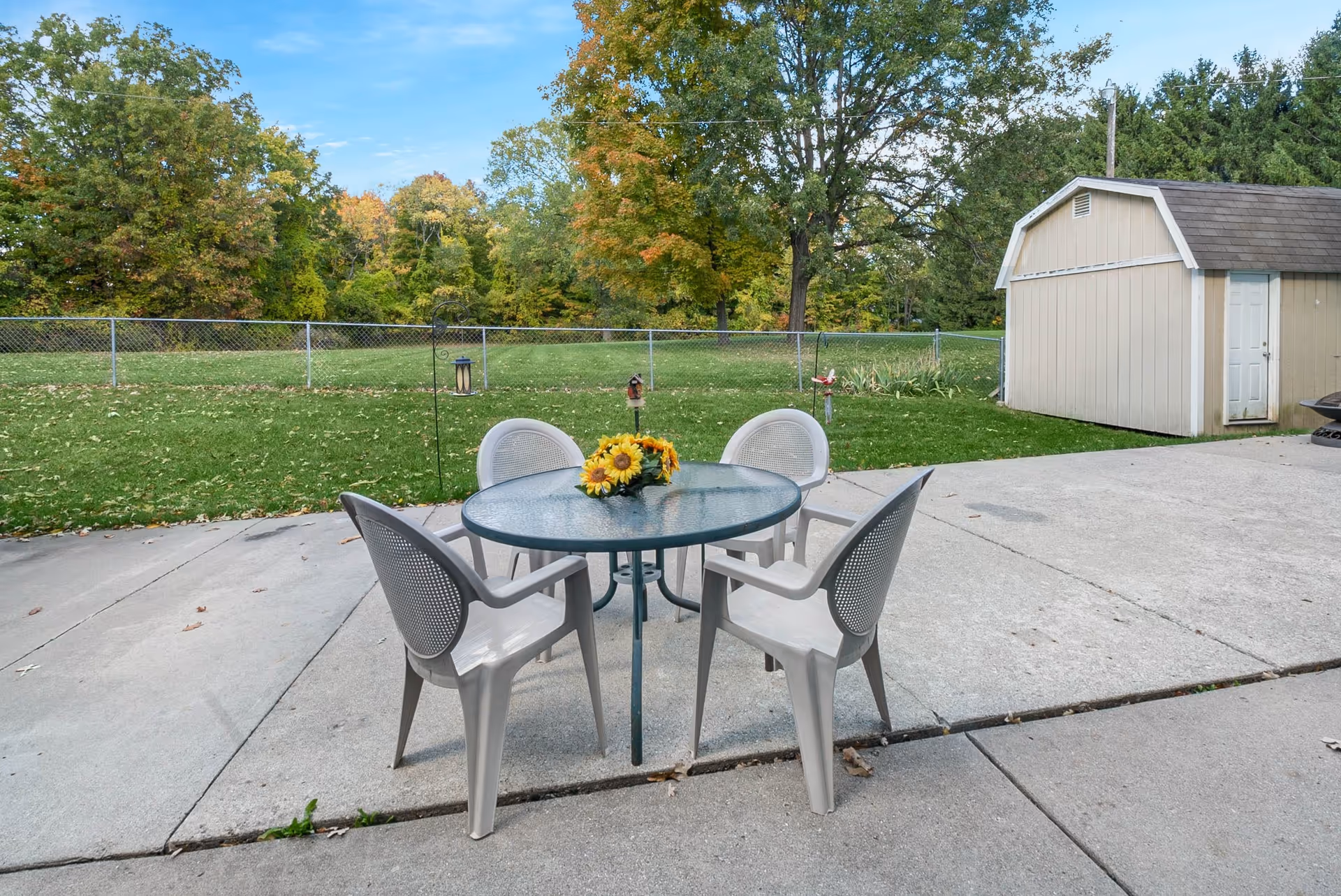 Outdoor patio area with a round glass table and four plastic chairs arranged around it. A bouquet of sunflowers is placed on the table. In the background, there is a green lawn, a chain-link fence, trees with autumn foliage, and a beige shed with a white door.