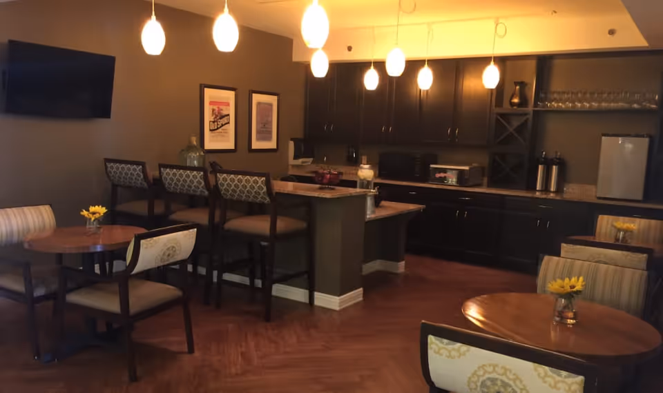 A cozy dining and kitchen area in a senior living facility featuring round wooden tables with chairs, a countertop bar with high chairs, dark cabinetry, a small refrigerator, microwave, and pendant lighting hanging from the ceiling. The walls are decorated with framed artwork and a mounted flat-screen TV.