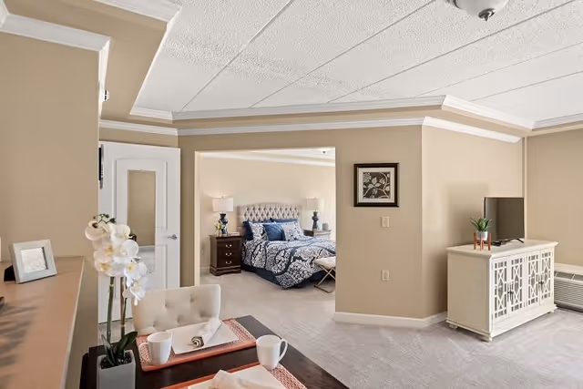 View of a cozy living area with a beige armchair and a small table set with two cups and a napkin. The room has beige walls and carpeted floors. In the background, there is a bedroom with a tufted headboard bed, blue and white bedding, two bedside tables with lamps, and a framed picture on the wall. A white TV stand with a flat-screen TV and a small plant is also visible.