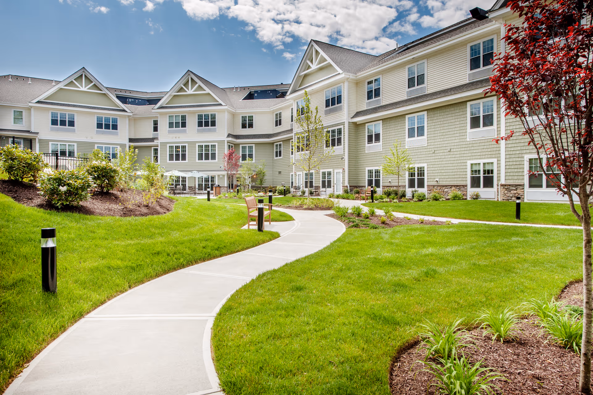 A landscaped outdoor garden area with a winding concrete pathway, green grass, small trees, bushes, and benches, adjacent to a multi-story senior living facility building under a partly cloudy sky.