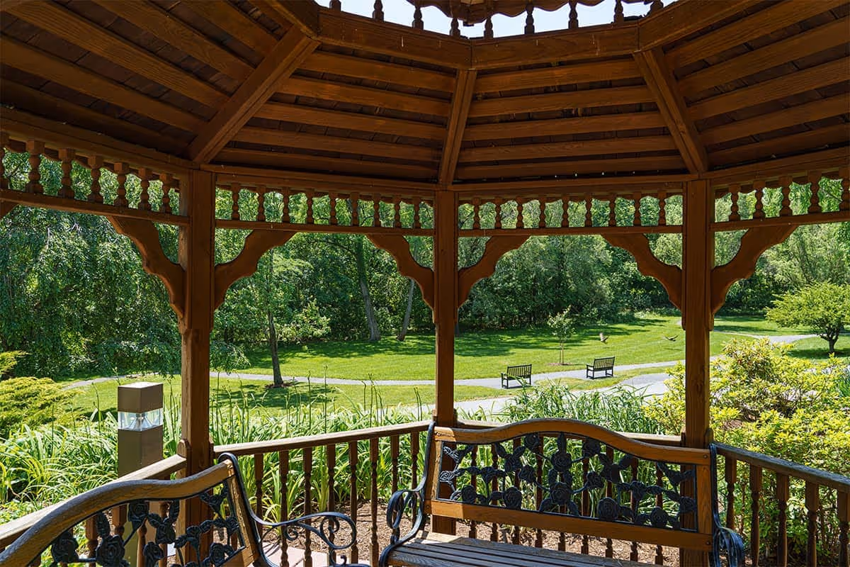 View from inside a wooden gazebo with decorative railings and a wooden roof, overlooking a green garden with trees, bushes, a walking path, and two benches in the distance.