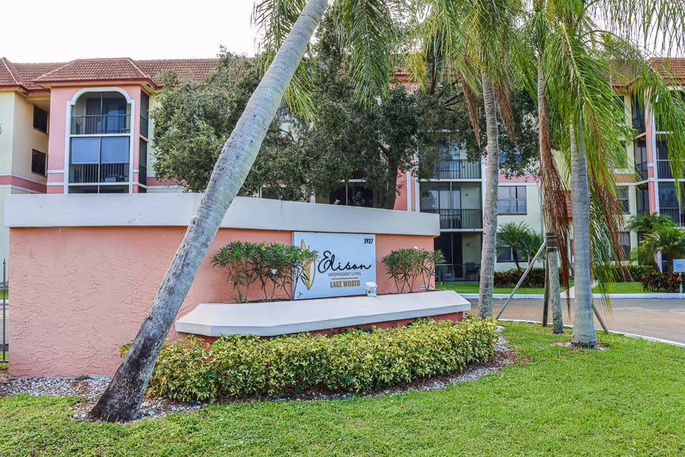Exterior view of Elison Independent Living of Lake Worth facility with a pink and white sign surrounded by greenery and palm trees in front of a multi-story building with balconies and windows.