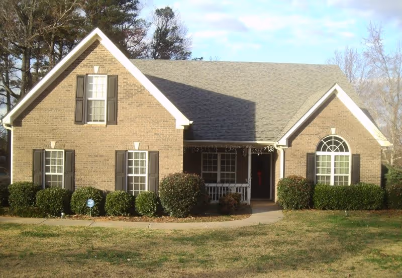 Front exterior view of a single-story brick house with a gray shingled roof, multiple windows with black shutters, a small covered porch with white railing, and neatly trimmed bushes along the front. The lawn is partially green and brown, and there are trees in the background under a partly cloudy sky.