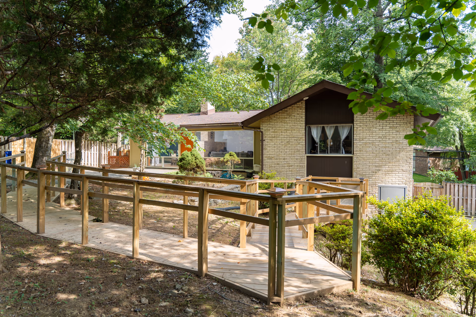 A single-story brick building surrounded by trees and greenery with a wooden ramp leading up to the entrance, providing accessible access. The building has a large window with white curtains and a brown section under the roof peak.