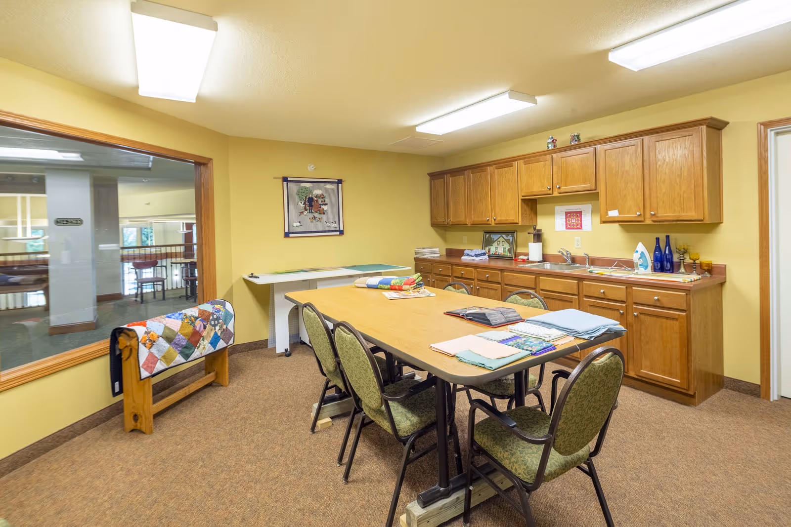 A craft room with a large table surrounded by six chairs, various folded fabrics on the table, wooden cabinets with a countertop and sink along one wall, an ironing board, and a quilt rack with a colorful quilt. The walls are painted yellow and there is a large window looking into another room.