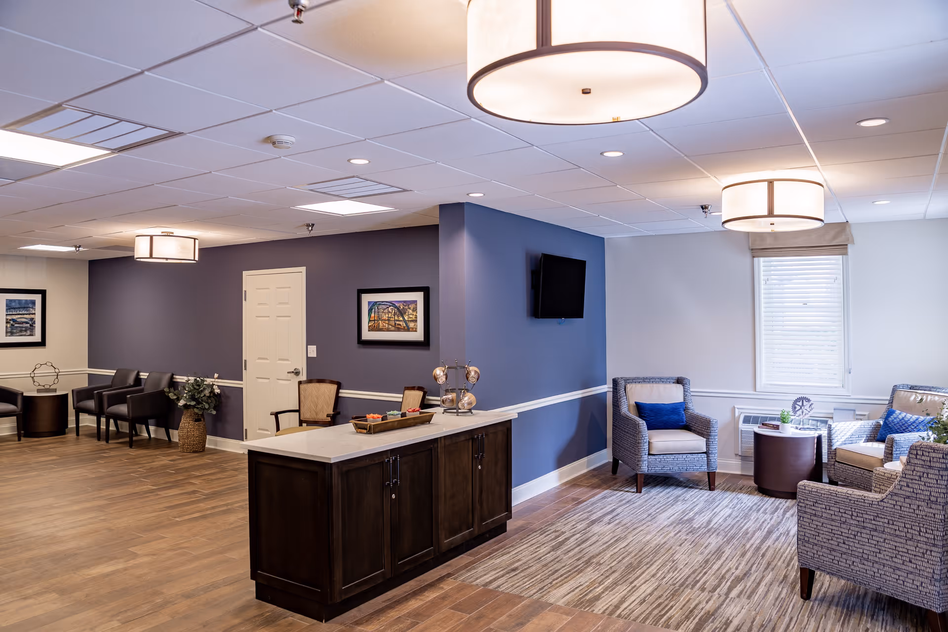 A spacious senior living facility common area with wooden flooring and a mix of dark and light blue walls. The room features a central island with a tray of snacks, several armchairs with blue cushions arranged around small tables, framed artwork on the walls, a wall-mounted TV, and large ceiling lights providing warm illumination.