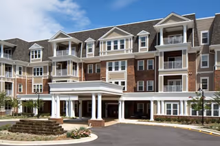 Exterior view of a multi-story senior living facility with a covered entrance, balconies, and a circular driveway with landscaped flower beds.