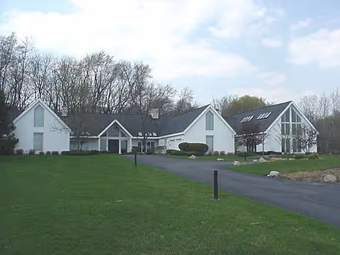 White single-story building with pitched roofs, a paved driveway and a large lawn with trees in the background.