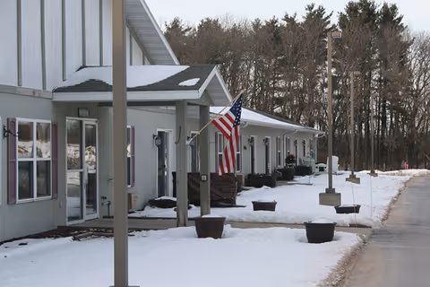 Exterior view of a single-story assisted living facility building in winter with snow covering the ground. The building has multiple doors and windows, an American flag mounted near one entrance, and several large planters along the walkway. Trees are visible in the background.