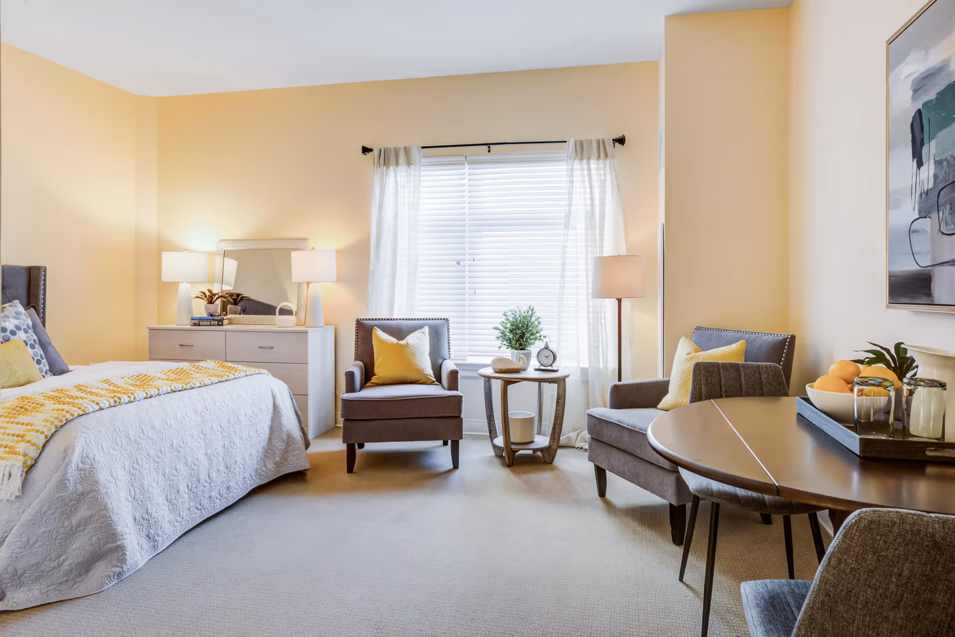A bright and cozy bedroom in a senior living facility featuring a neatly made bed with white and yellow bedding, two gray armchairs with yellow pillows, a small round wooden table with a plant and clock, a dresser with two lamps and a mirror, and a round dining table with chairs and a tray holding fruit and glasses. The room has beige walls and carpet, and a window with white blinds and sheer curtains.