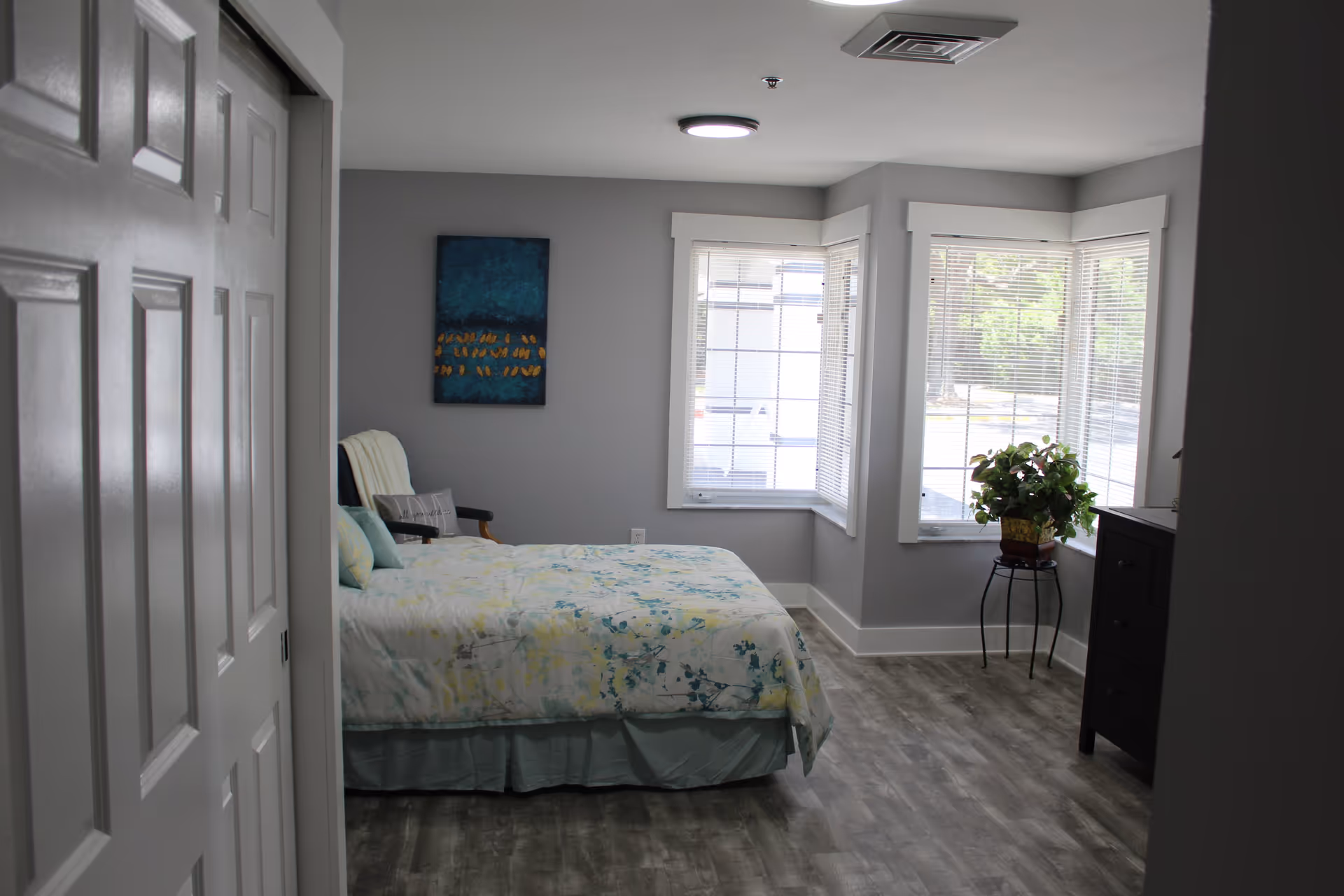 A bright bedroom with a bed covered in a light-colored floral bedspread, a chair with a pillow, a painting on the wall, large windows with blinds letting in natural light, a small table with a potted plant, and a dark dresser.