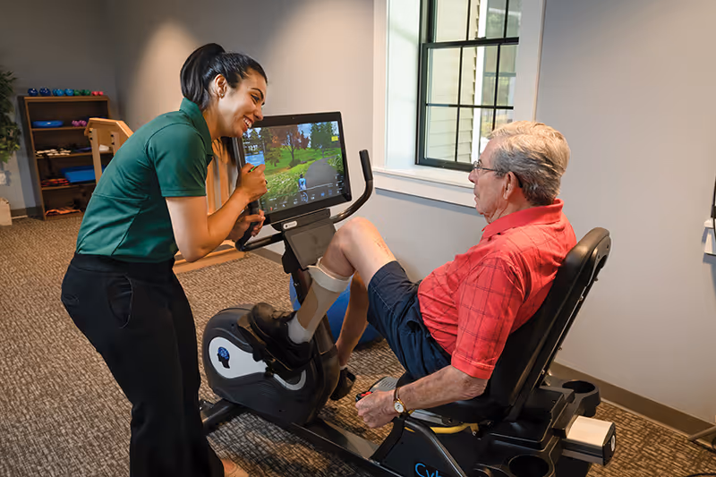 An elderly man in a red shirt and black shorts is seated on a recumbent exercise bike, pedaling while looking at a screen displaying a virtual cycling path. A woman in a green shirt and black pants is standing next to him, smiling and engaging with him. The room has carpeted flooring, a window, and shelves with exercise equipment in the background.