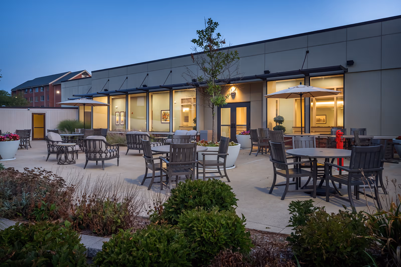 Outdoor courtyard with multiple tables, chairs, umbrellas and planters in front of a single-story building at dusk.