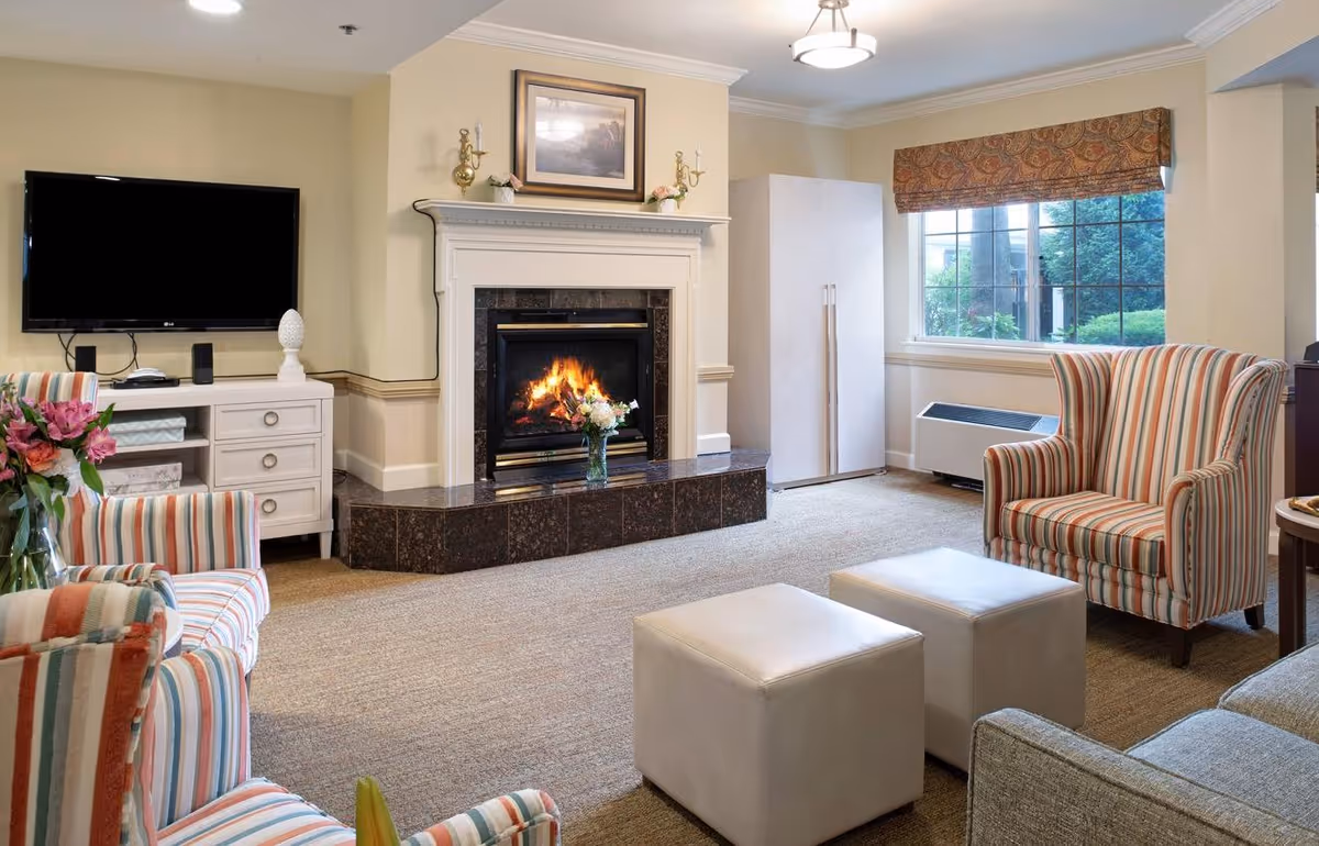A cozy living room with a lit fireplace in the center, flanked by a white cabinet with a TV mounted above it on the left and a large window with a patterned valance on the right. The room features striped armchairs, two white ottomans, a gray sofa, and a vase of flowers on a side table. The walls are painted light beige and the floor is carpeted.