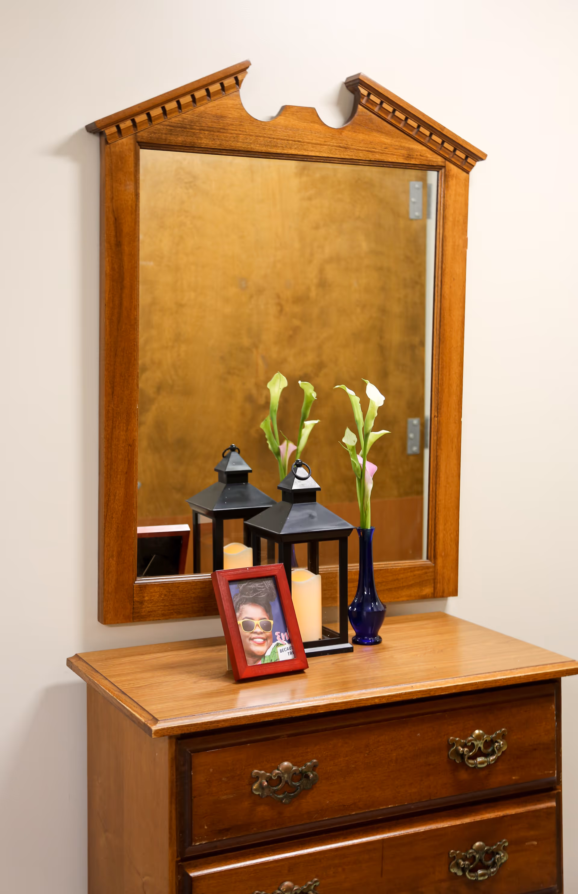 Wooden dresser topped with a framed photo, decorative lanterns, and a vase of flowers in front of a wall-mounted mirror.