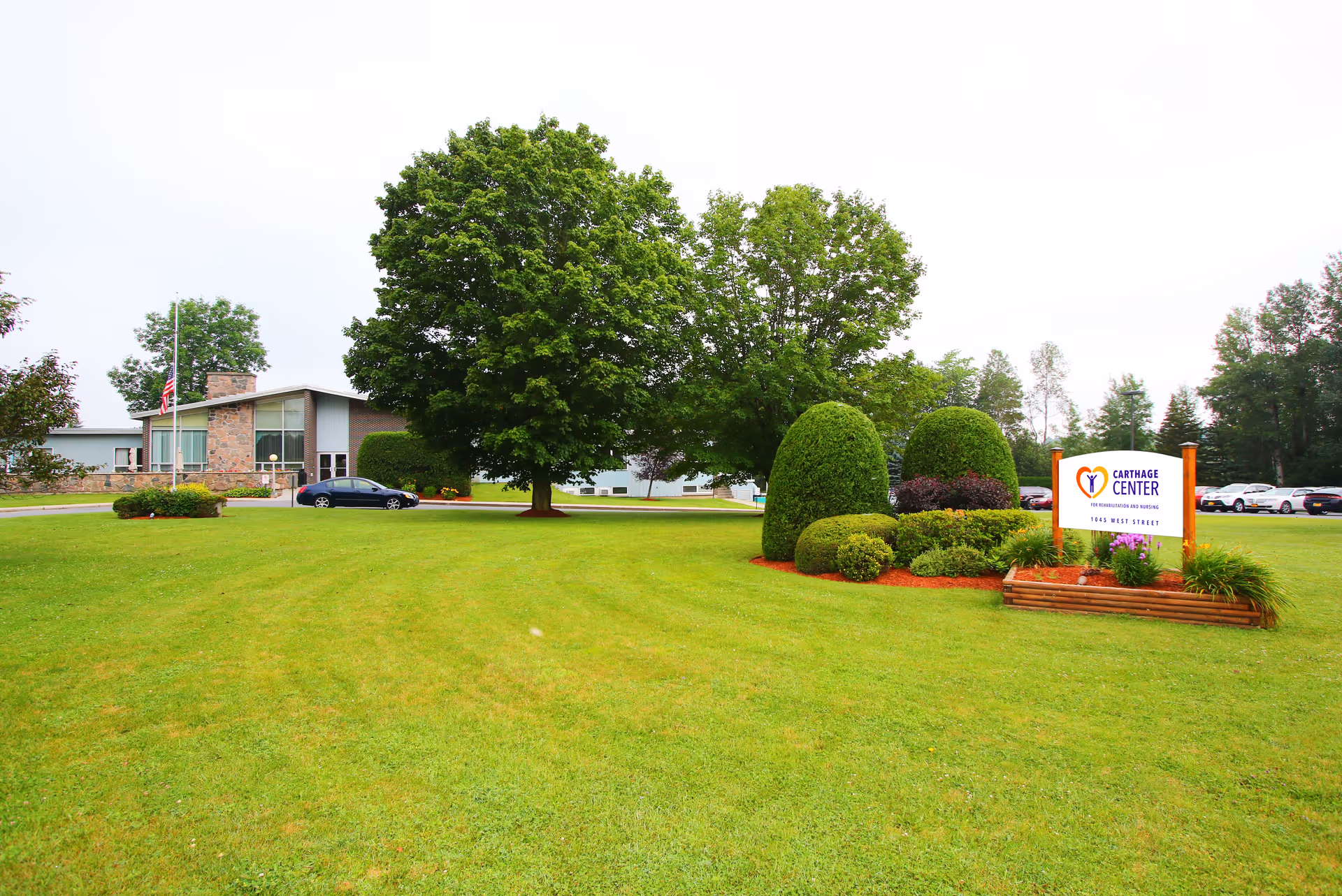 A well-maintained lawn with neatly trimmed bushes and trees in front of a single-story building. There is a sign on the right side that reads 'Carthage Center' with the address 1803 West Street. Several parked cars are visible in the background near the building.