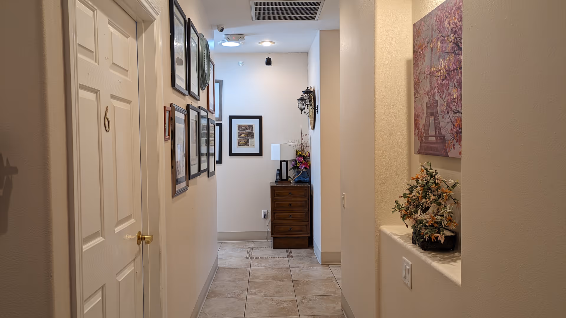 A well-lit hallway in a senior living facility with beige walls and tiled floor. On the left side, there is a white door marked with the number 6 and several framed pictures hanging on the wall. At the end of the hallway, there is a wooden chest of drawers with a lamp and a flower arrangement on top. On the right side, there is a recessed wall niche with a flower pot and a painting of the Eiffel Tower with cherry blossoms above it.
