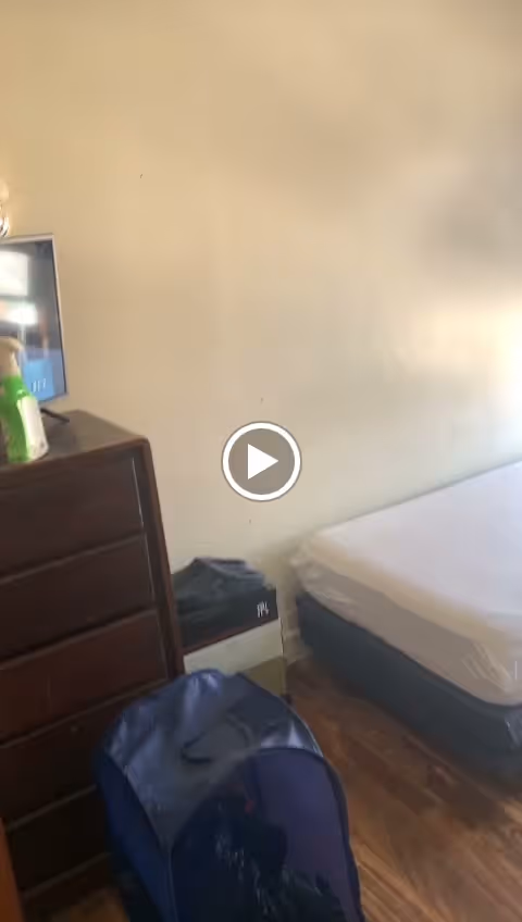 A simple bedroom with a mattress on a bed frame, a wooden dresser with a TV on top, and a blue laundry hamper on a wooden floor. The walls are plain and light-colored.