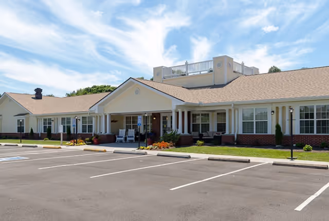 Single-story senior living building with a covered entrance, porch seating, landscaping and empty parking spaces under a blue sky.
