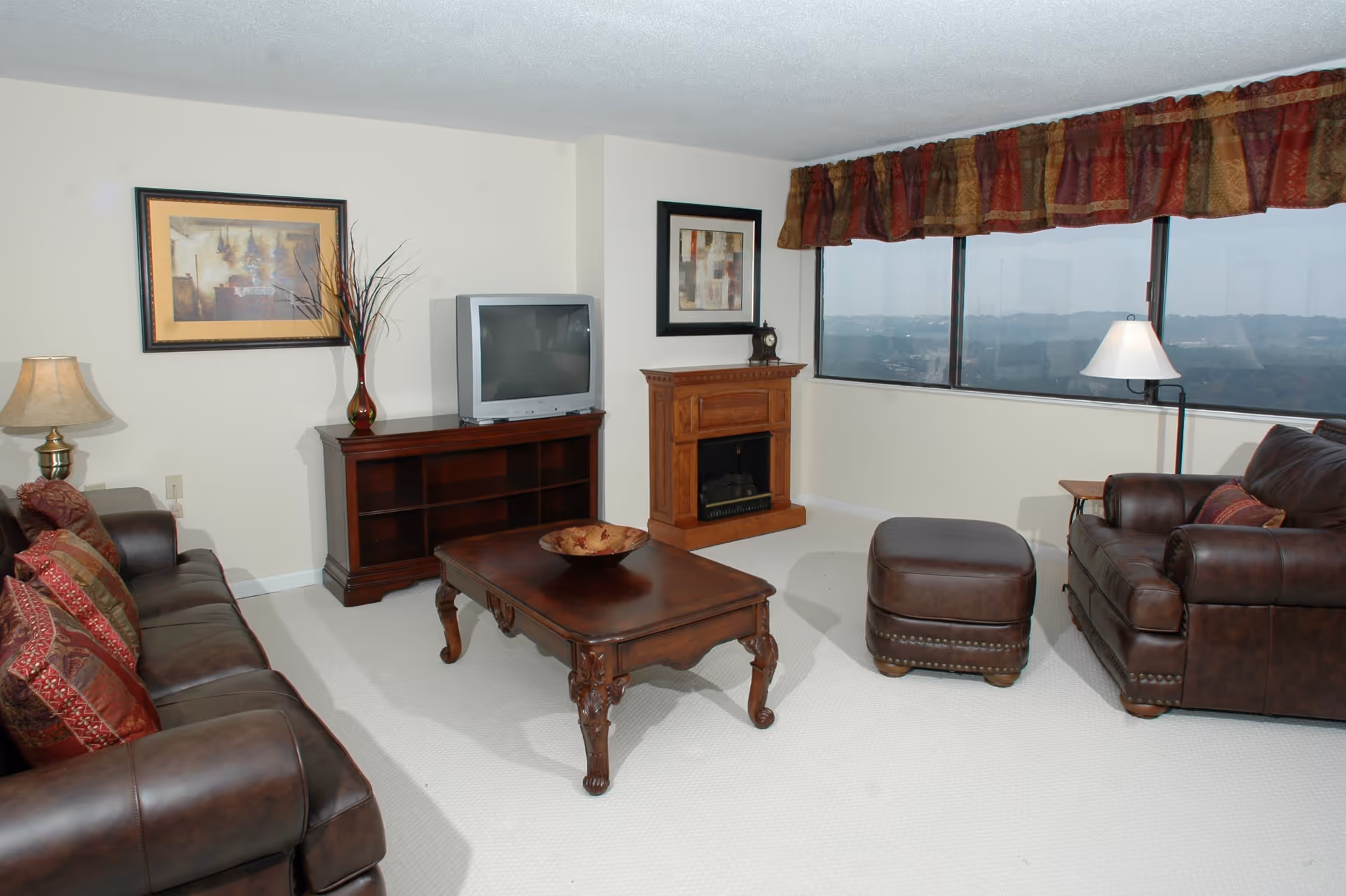 A living room with brown leather sofa and armchair, a wooden coffee table with a decorative bowl, a wooden TV stand with an older style television, a small wooden fireplace, a floor lamp, and large windows with a view outside. The room has light-colored walls and carpet, with framed artwork and a vase with decorative branches.