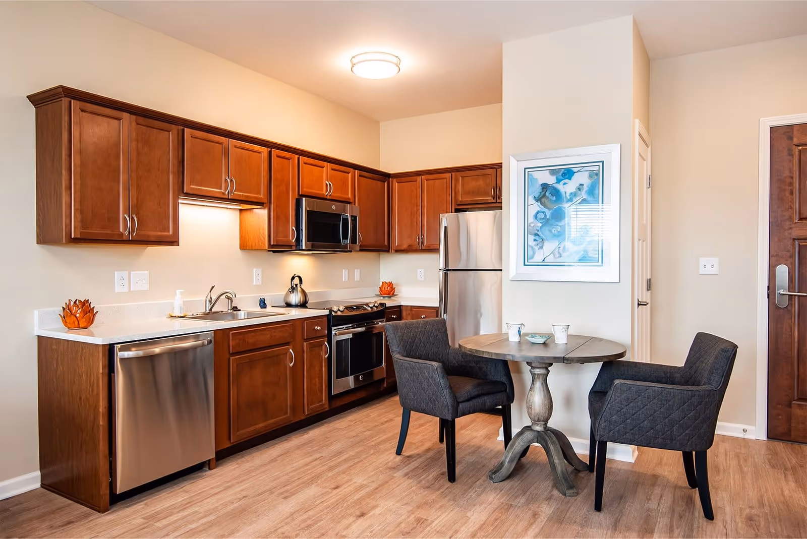 A modern kitchen with wooden cabinets, stainless steel appliances including a dishwasher, oven, microwave, and refrigerator. There is a small round wooden table with two dark upholstered chairs. A framed artwork with blue flowers hangs on the wall next to the table. The floor is light wood, and the walls are painted beige.
