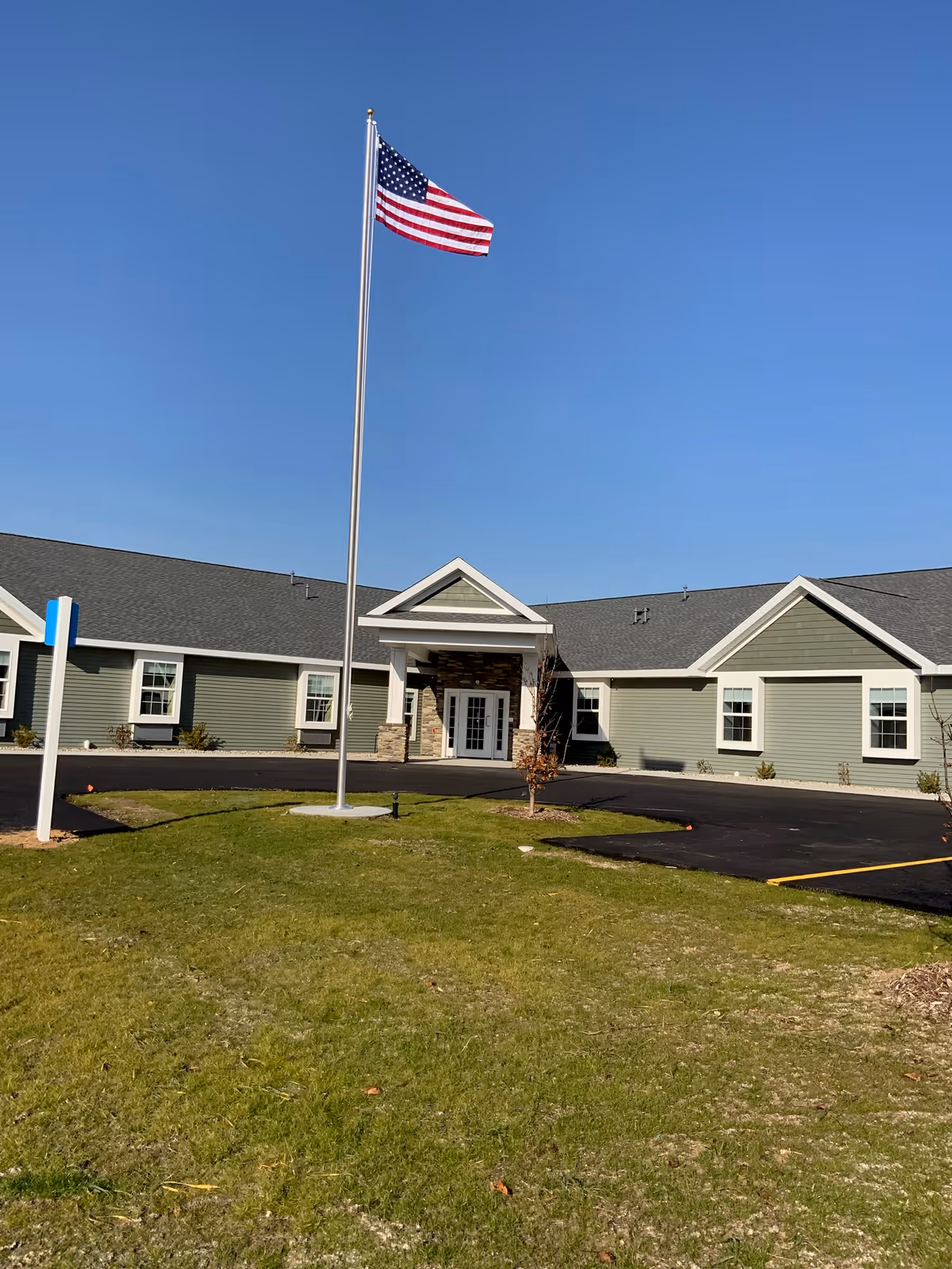 Exterior view of a single-story assisted living facility building with green siding and white trim under a clear blue sky. An American flag is flying on a tall flagpole in front of the building, which has a paved driveway and a small grassy area.