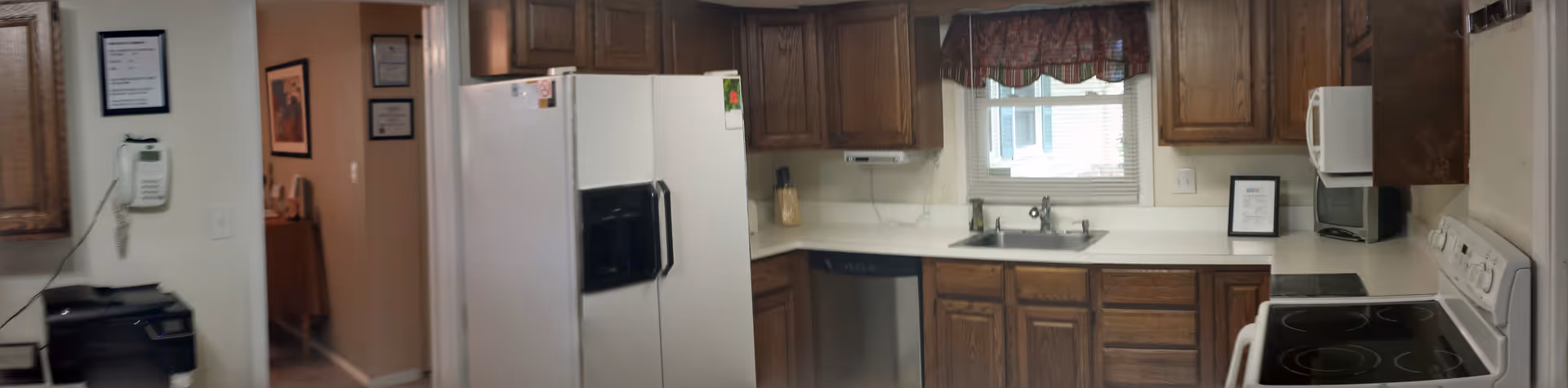 Interior view of a kitchen with wooden cabinets, a white refrigerator, a dishwasher, a microwave, an electric stove, and a sink under a window with a decorative valance. A wall-mounted phone and a printer are visible on the left side near the entrance to another room.