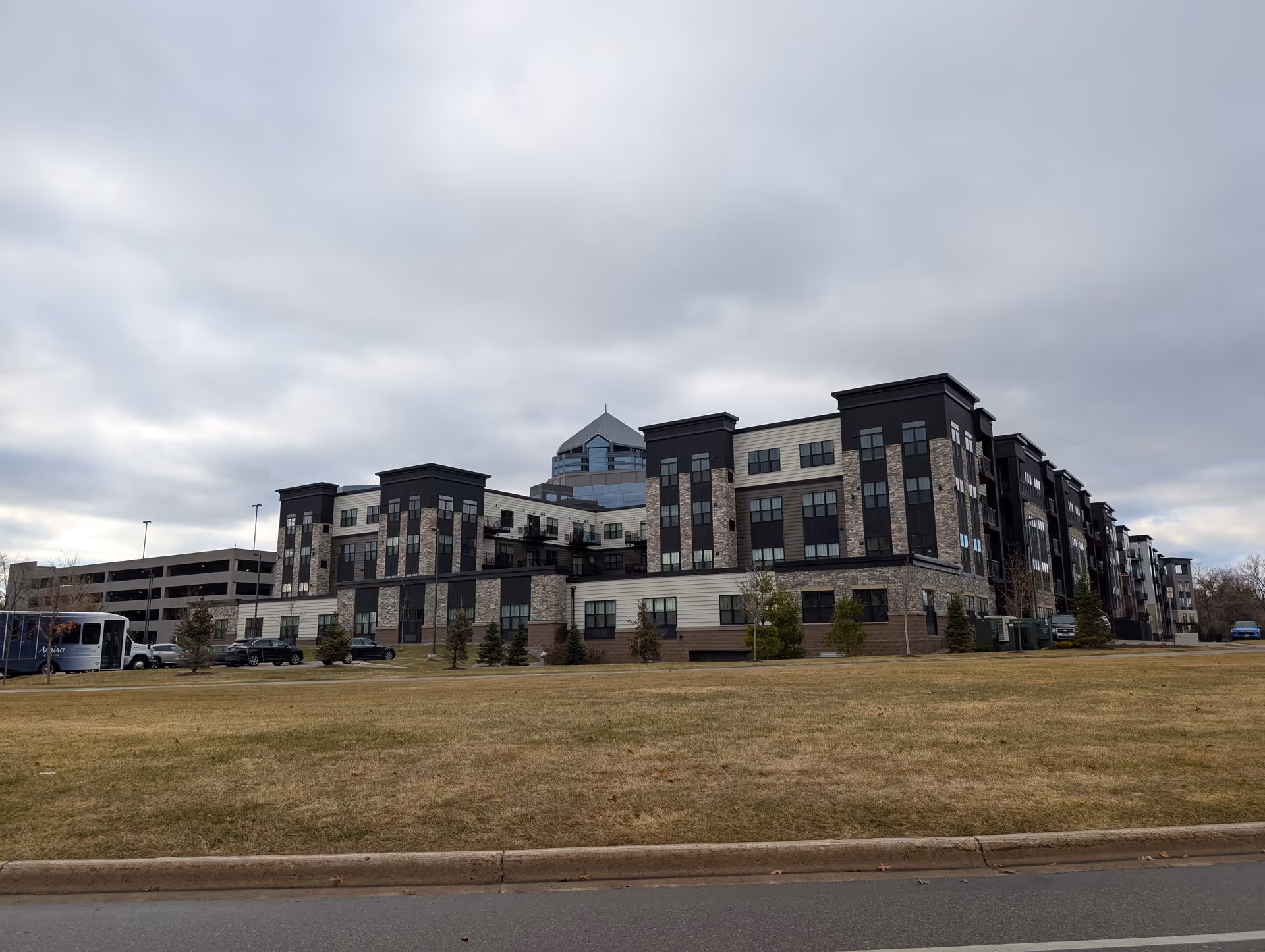 Exterior view of a large multi-story senior living facility building with a mix of dark and light-colored siding and stone accents, surrounded by a grassy area and a few small trees under a cloudy sky.