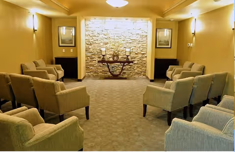 A cozy seating area with multiple beige armchairs arranged in two rows facing each other. The room has warm yellow lighting, a stone accent wall at the far end with a decorative table holding a vase and other decor items. Two framed pictures hang on the walls on either side of the stone wall.