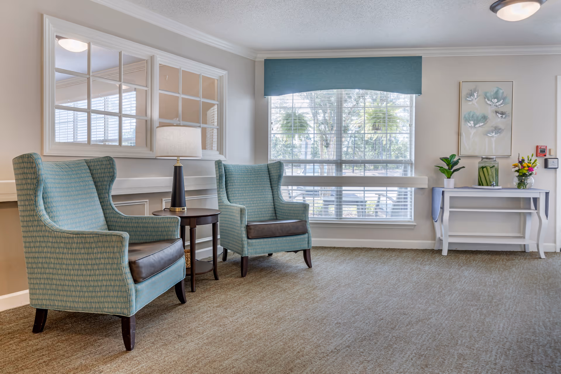A bright and cozy sitting area with two teal upholstered armchairs with dark brown leather seat cushions, separated by a small round wooden table with a lamp. Behind the chairs is a large window with white blinds and a teal valance, allowing natural light to fill the room. On the right side, there is a white console table decorated with a potted plant, a large glass jar with green contents, a vase of colorful flowers, and a framed floral artwork on the wall above. The room has beige walls, crown molding, and a beige carpeted floor.