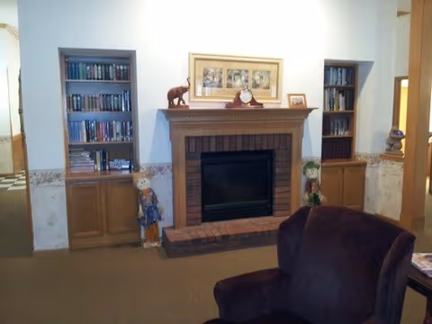 Cozy common room with a central brick fireplace flanked by built-in bookshelves, decorative items, and an upholstered armchair in the foreground.