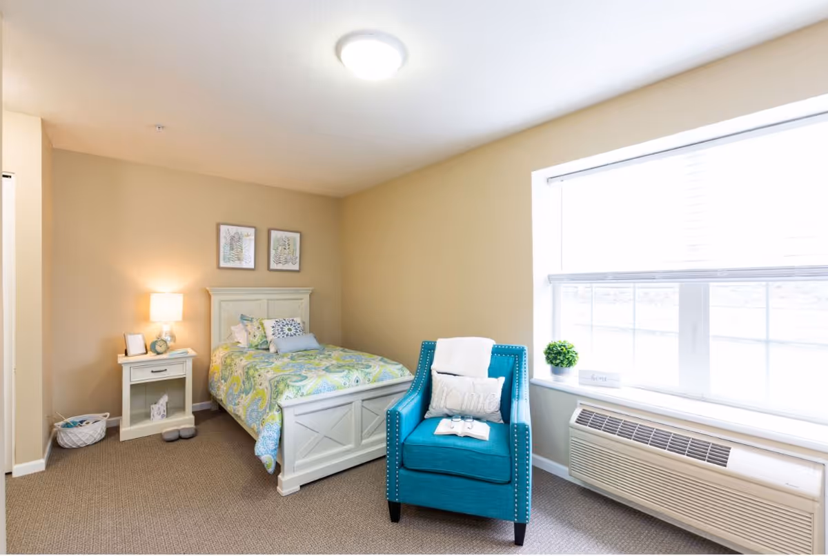 A bright bedroom with a single bed featuring a green and blue patterned bedspread, a white nightstand with a lamp and clock, a teal armchair with pillows and a white throw blanket, a large window with blinds, and a small potted plant on the windowsill.