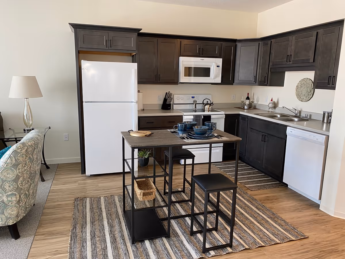 Open kitchen with dark cabinets, white appliances, a small island with stools, and a partial view of the living area.