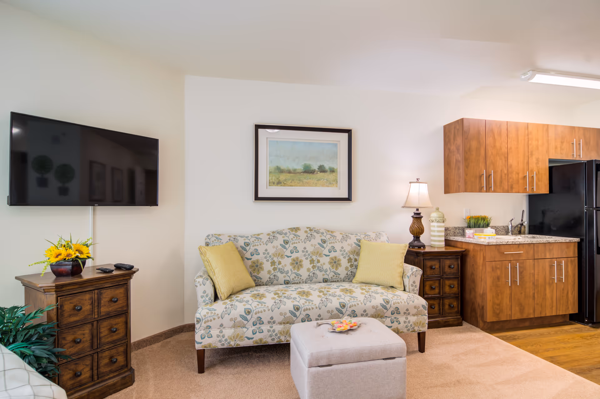 A cozy living area in a retirement facility featuring a floral patterned sofa with two yellow cushions, a small wooden side table with a lamp and decorative items, a wall-mounted flat-screen TV above a wooden cabinet with a vase of sunflowers, and a kitchenette with wooden cabinets and a black refrigerator.