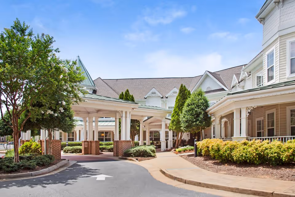 Exterior view of a senior living facility with a covered entrance supported by white columns and brick bases, surrounded by well-maintained landscaping including trees and shrubs under a blue sky.