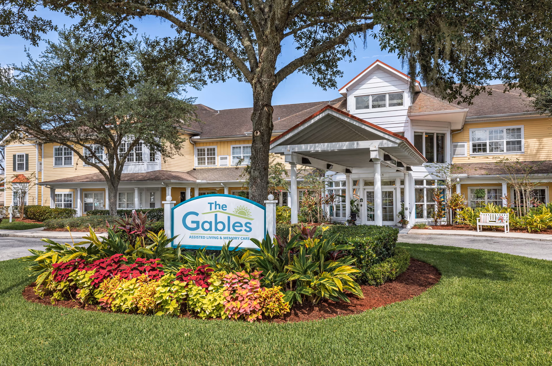 Front exterior view of The Gables of Jacksonville assisted living and memory care facility with a landscaped garden featuring colorful plants and a large tree in front of the building entrance.