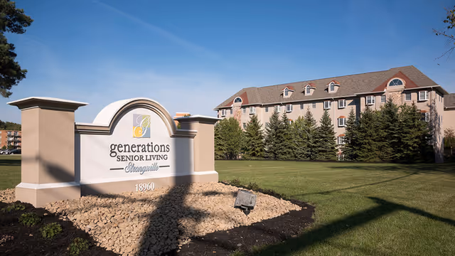 Exterior view of Generations Senior Living of Strongsville building with a large sign in the foreground displaying the facility name and address, surrounded by a well-maintained lawn and trees under a clear blue sky.