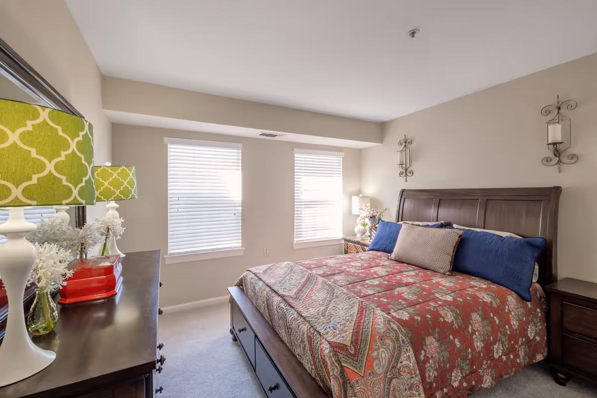 A bedroom with a large wooden bed featuring a red floral patterned bedspread and multiple pillows in blue and beige. There are two windows with white blinds letting in natural light. On the left side, there is a dark wooden dresser with a green patterned lamp, white flowers in a vase, and a red box. On the right side of the bed, there is a nightstand with a lamp and flowers. The walls are beige with two decorative wall sconces above the bed.